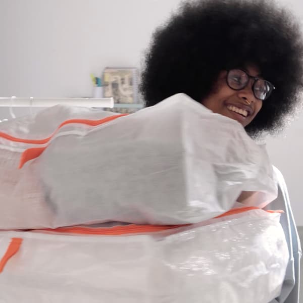 Une jeune femme souriante dans une pièce claire portant deux sacs de rangement PÄRKLA blancs avec des détails orange.