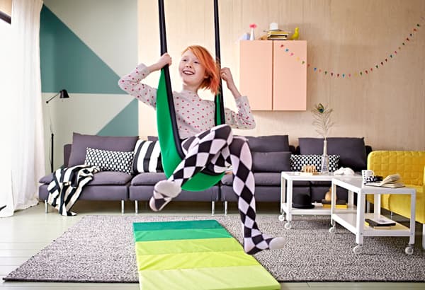 Une jeune fille assise dans une balançoire, dans le salon, avec un tapis de gymnastique en dessous.