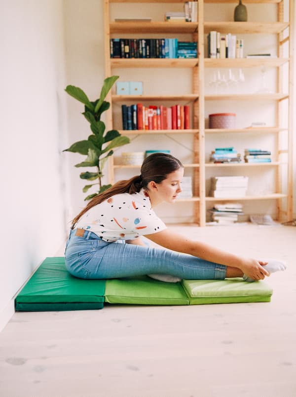 Une jeune fille fait des étirements, assise sur un tapis de gymnastique pliant vert PLUFSIG, avec une plante et une étagère IVAR à l’arrière-plan.