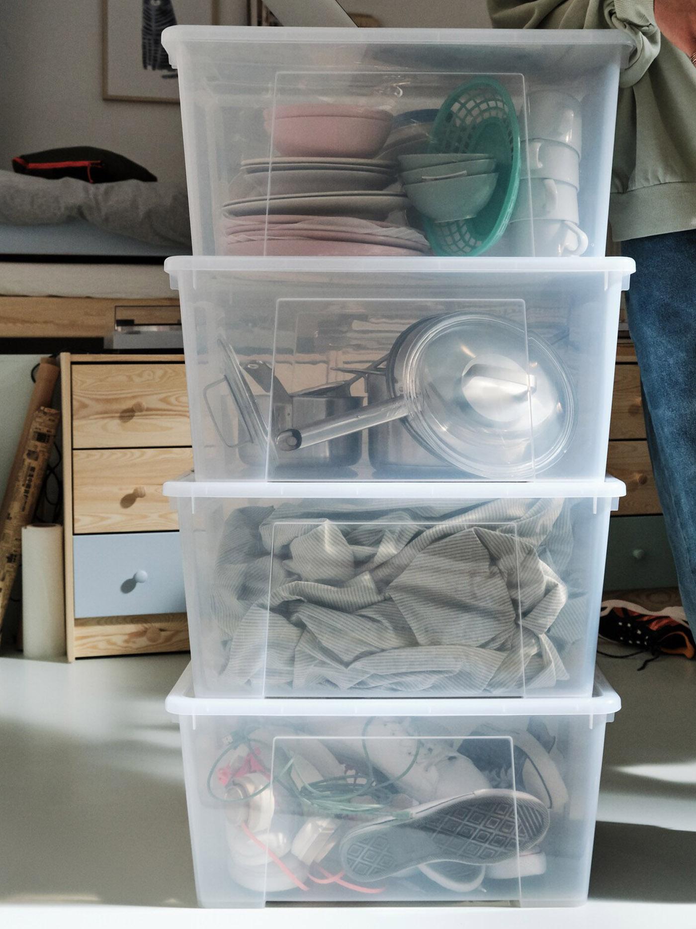 Une jeune personne à côté de quatre boîtes SAMLA 45 l transparentes empilées les unes sur les autres, contenant des chaussures et des ustensiles de cuisine.