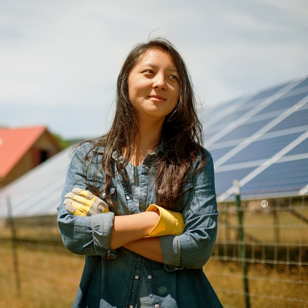 Une jeune personne vêtue d’une chemise bleue en jean et de gants de travail se tient debout, les bras croisés, devant un ensemble de panneaux solaires.