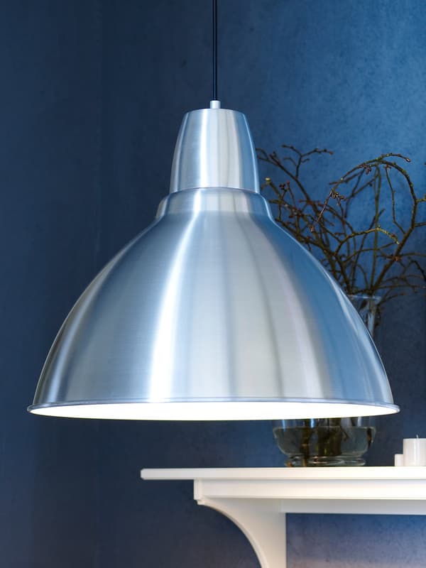 Une lampe FOTO suspendue dans une pièce aux murs bleus avec un manteau de cheminée en bois blanc, sur lequel se trouvent des branches dans un vase.