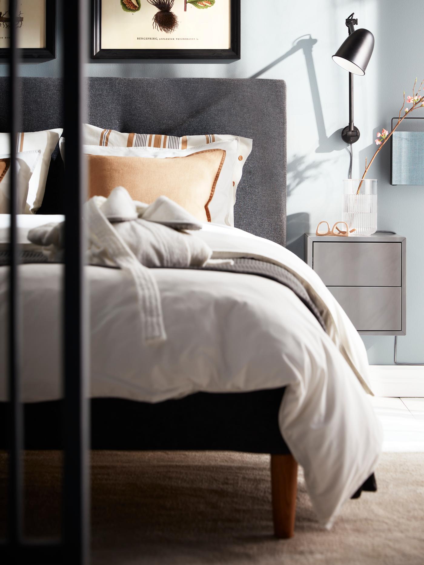 Une lampe murale SKURUP noire, une armoire EKET gris foncé et une enceinte Bluetooth fixée à un mur à côté d’un lit IDANÄS gris foncé.