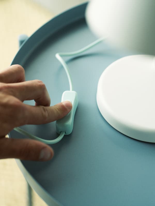 Une main actionne l’interrupteur d’une lampe de table blanche posée sur une table bleu clair.
