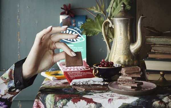 Une main attrapant un carré de chocolat et une table décorée de barres de chocolat, de livres et de fruits.