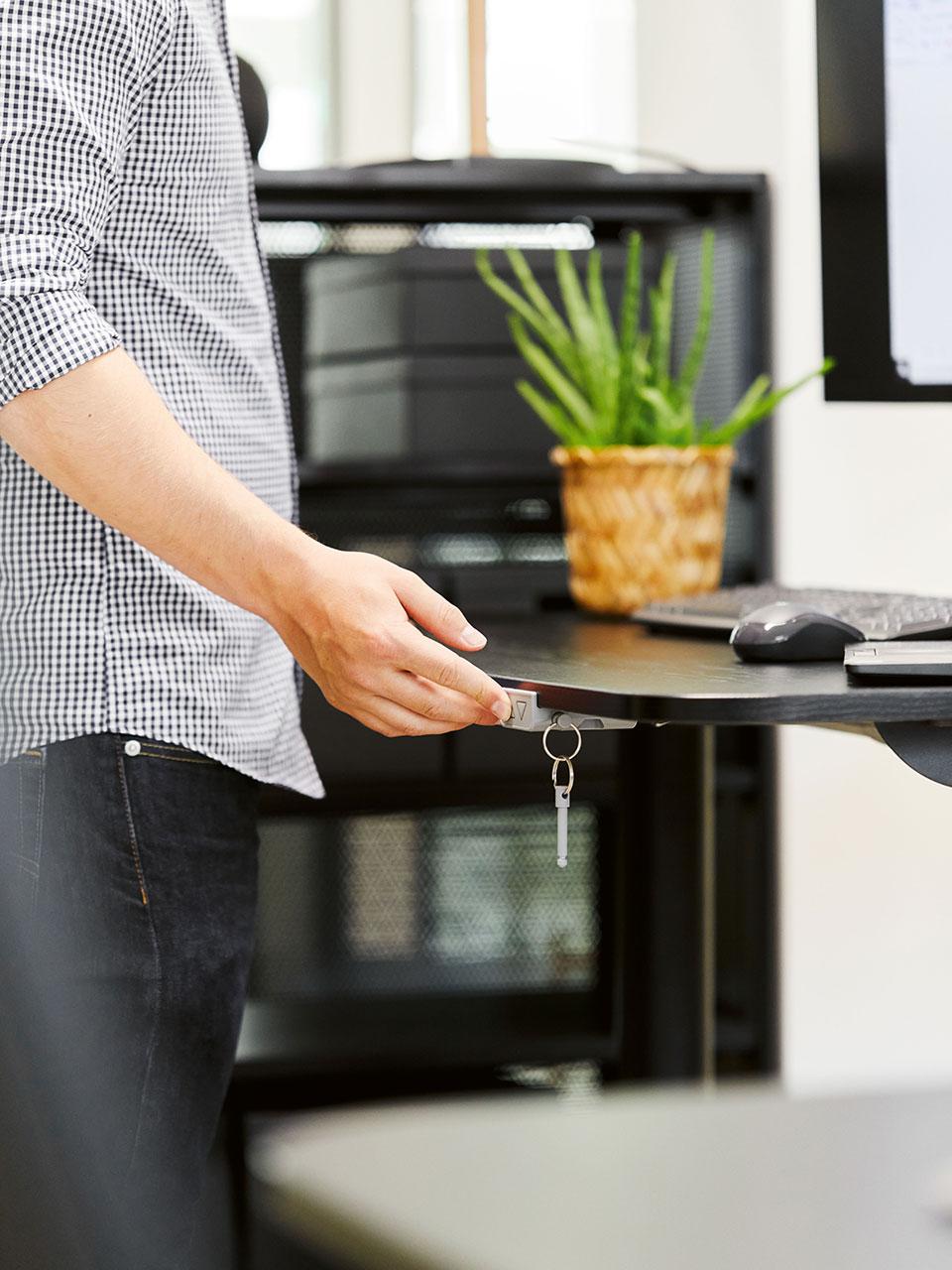 Une main manipule un bureau noir à réglage électrique, sur lequel se trouvent une souris, un clavier et une petite plante en pot dans un cache-pot en rotin. 