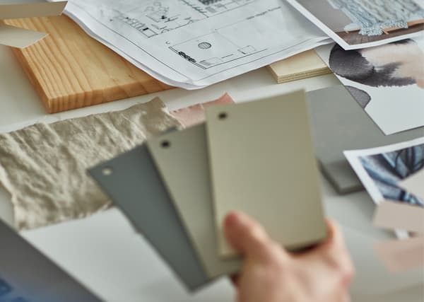 Une main tenant des échantillons de couleur dans les tons gris et beige, au-dessus d’un bureau couvert de plans, photos et échantillons.