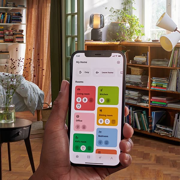 Une main tenant l'application IKEA Home Smart devant un salon affichant un écran avec différentes options.