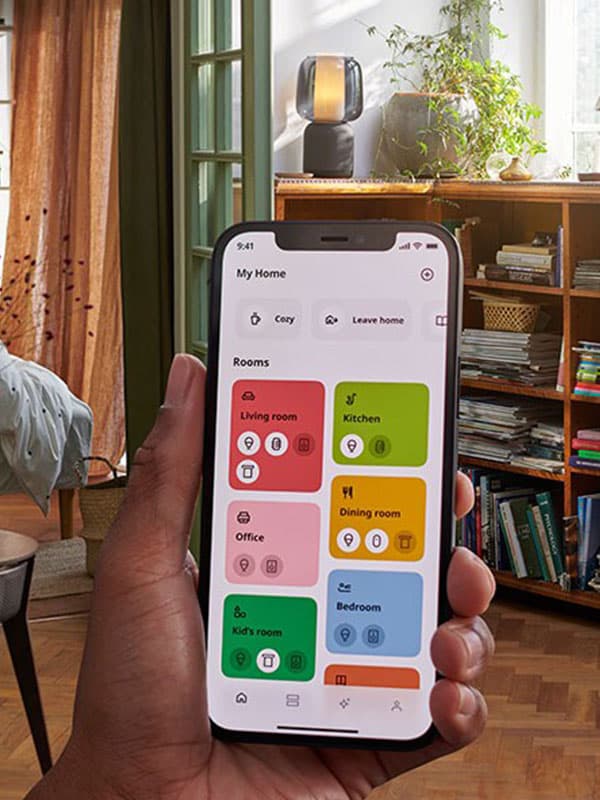 Une main tenant l'application IKEA Home Smart devant un salon affichant un écran avec différentes options.