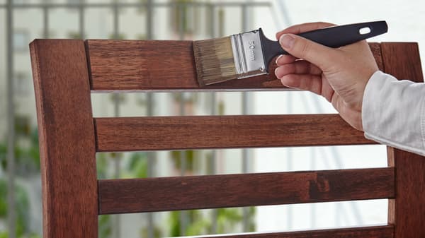 Une main tenant un pinceau et lasurant le dos d’une chaise d’extérieur en bois.