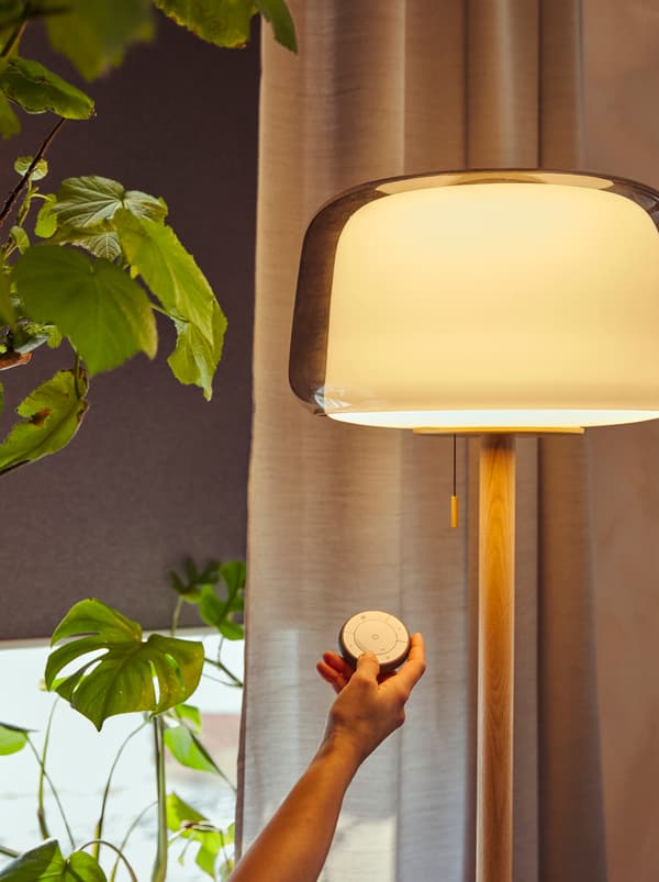 Une main tend une télécommande TRÅDFRI vers un lampadaire EVEDAL allumé. Derrière se trouve une fenêtre devant laquelle un store TRETUR est partiellement abaissé.