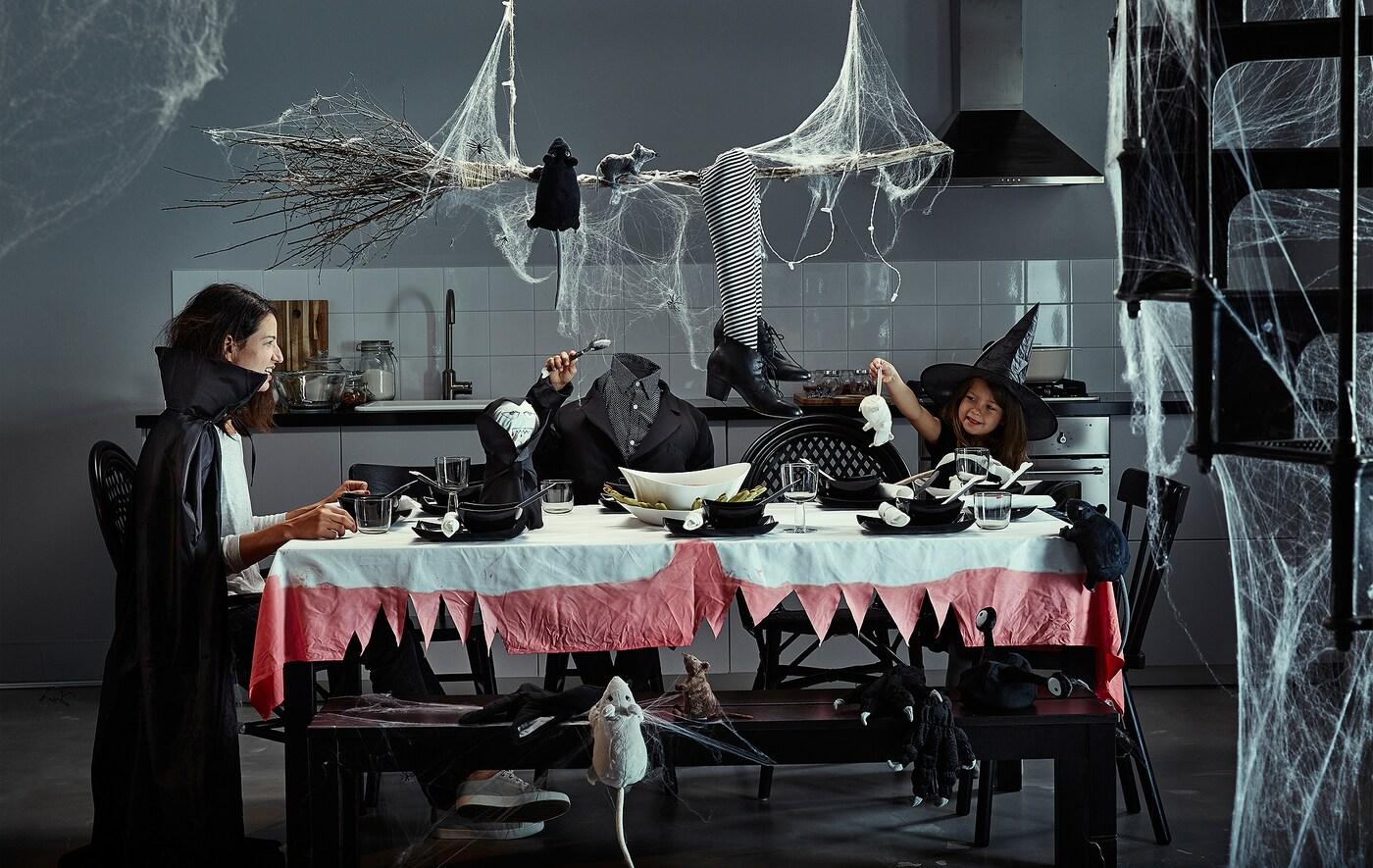 Une mère et ses enfants assis à une table décorée pour Halloween avec des toiles d’araignée, des costumes et des décorations effrayantes.