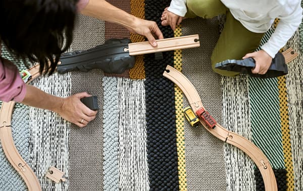 Une mère et son enfant construisent ensemble un circuit en bois sur un tapis tissé avec différentes rayures colorées.
