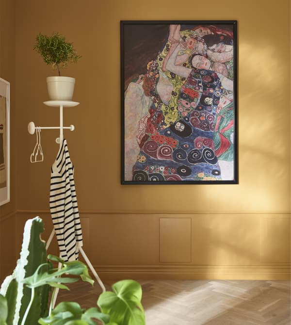 Une œuvre d’art sous cadre est accrochée à un mur ocre, avec un valet dans le coin.