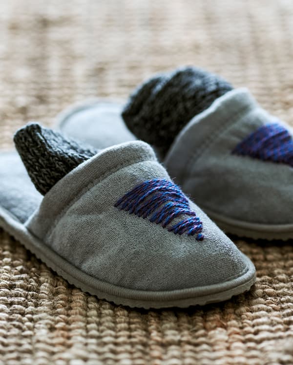 Une paire de pantoufles grises sur un tapis en jute, personnalisées avec du fil de laine bleu cousu sur le devant pour former un motif de sapin.
