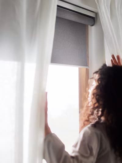 Une personne aux cheveux bouclés ouvrant des voilages blancs à côté d'une fenêtre avec un store à enrouleur gris, la lumière du soleil entrant dans la pièce.