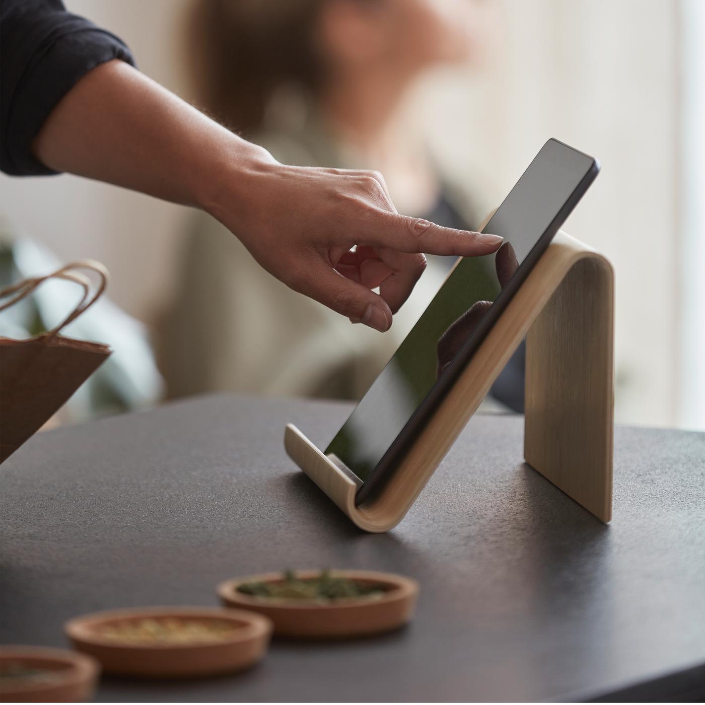 Une personne consulte une tablette posée sur un support pour tablette en bambou posé sur un plan de travail noir. Une autre personne se trouve près d’une fenêtre.