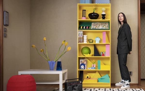 Une personne debout à côté d’une bibliothèque BILLY jaune vif avec des objets décoratifs sur les tablettes.
