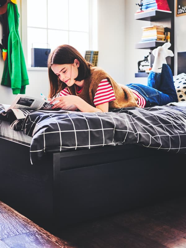 Une personne en train de lire allongée sur un lit MALM noir avec deux boîtes de rangement et garni d’une parure VITKLÖVER pour couette et oreiller.