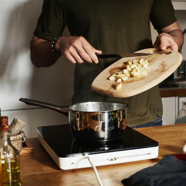 Une personne faisant glisser des pommes de terre émincées d’une planche à découper dans une casserole chromée, sur une table de cuisson à induction mobile TILLREDA.