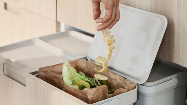 Une personne jette une épluchure de légume dans un sac à déchets alimentaires en papier brun. Ce dernier se trouve dans une solution de tri des déchets HÅLLBAR qui, elle, est installée dans un tiroir haut MAXIMERA.