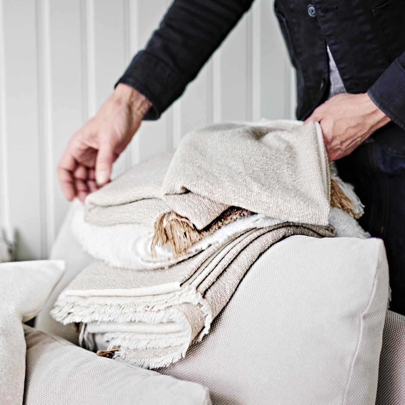 Une personne pliant un plaid GUNNEMOR gris-beige sur l’accoudoir d’un module d’angle SÖDERHAMN beige clair à 1 place.