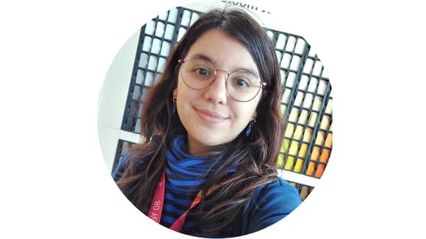 Une personne portant des lunettes et un haut bleu avec un badge est placée devant un arrière-plan composé de carrés colorés.