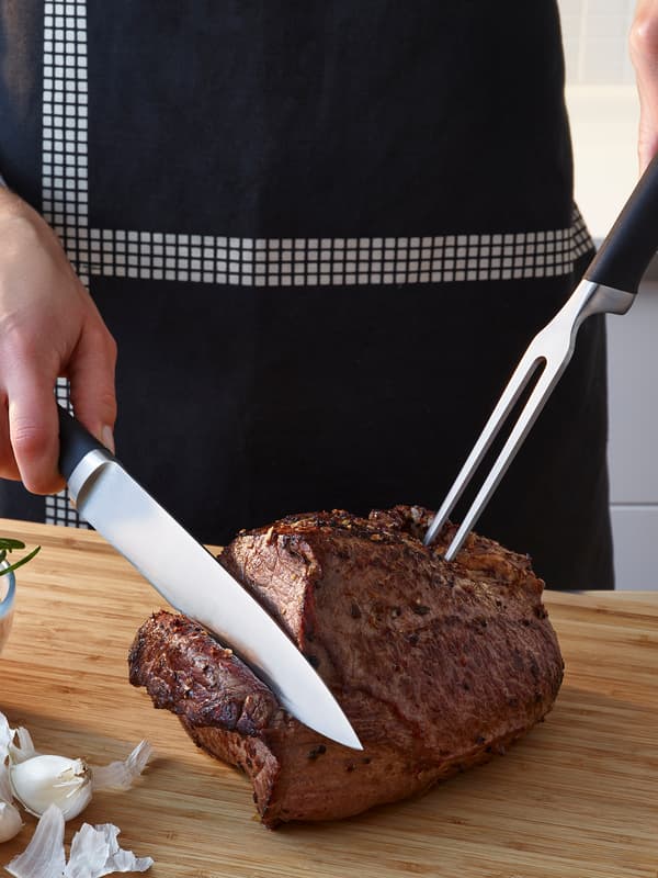 Une personne portant un tablier noir et blanc utilise une fourchette et un couteau à viande à manche noir pour couper de la viande.