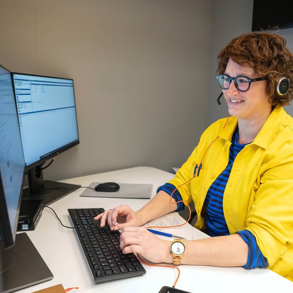 Une personne porte une veste jaune avec le logo IKEA et travaille à un bureau avec deux écrans. Elle utilise un clavier et porte un casque.