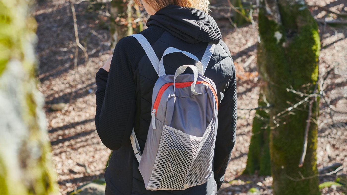 Une personne qui marche près d’un flanc de colline partiellement couvert par des arbres, portant une veste et un sac à dos PIVRING gris pâle.
