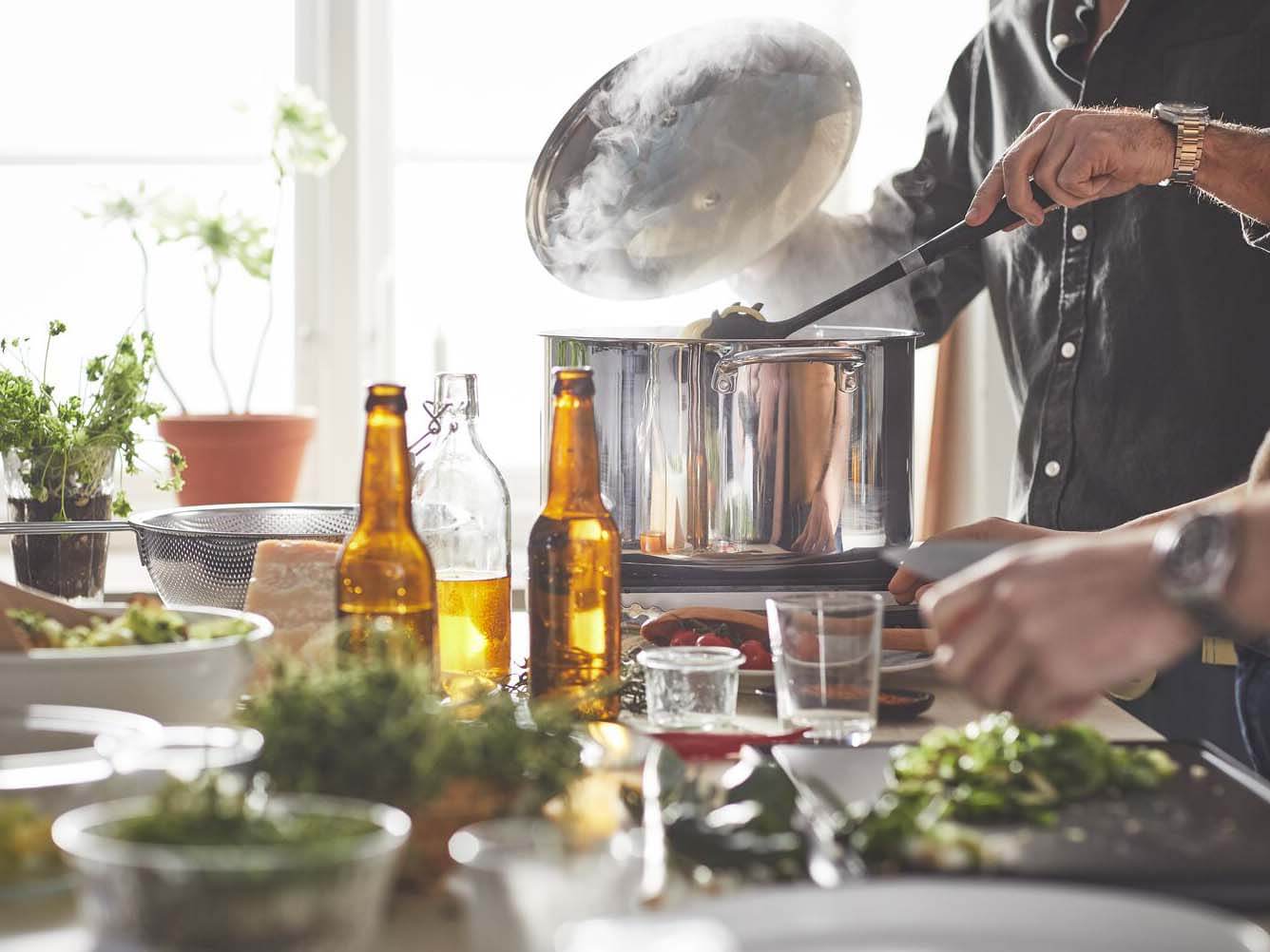Une personne qui tient un couvercle au-dessus d’une casserole bouillante tout en en remuant le contenu avec l’autre main devant une fenêtre ensoleillée. Des bouteilles, des herbes et d’autres ustensiles de cuisine sont sur le comptoir à côté.