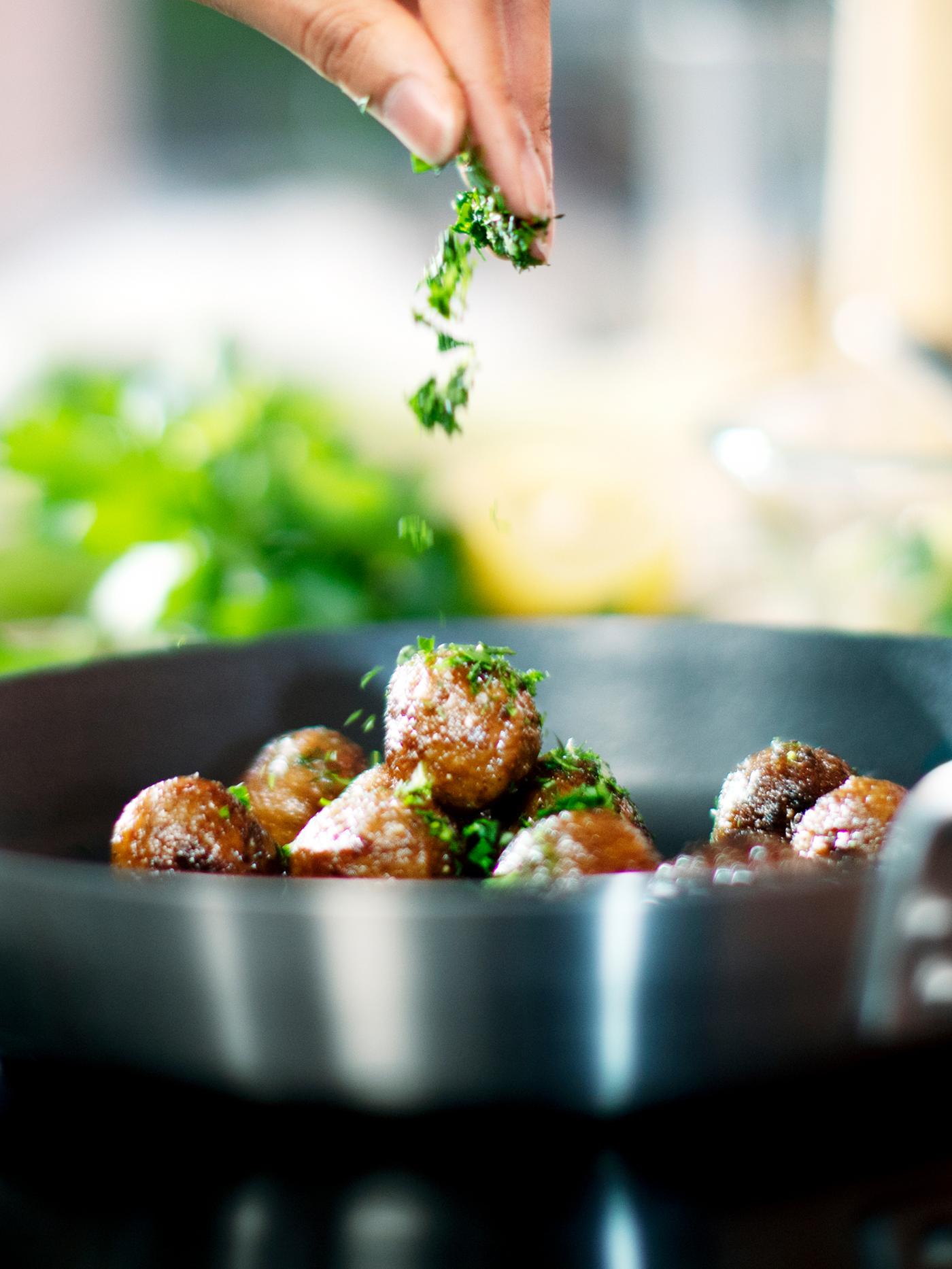 Une personne saupoudre des herbes vertes hachées sur des boulettes végétales HUVUDROLL disposées dans une poêle IKEA 365+ placée sur une table de cuisson.