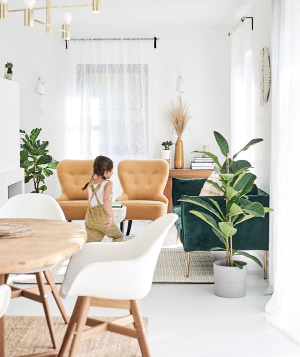 Une petite fille court dans un espace salle à manger/séjour décloisonné avec un canapé en velours vert, des fauteuils moutarde et des chaises de salle à manger blanches. 