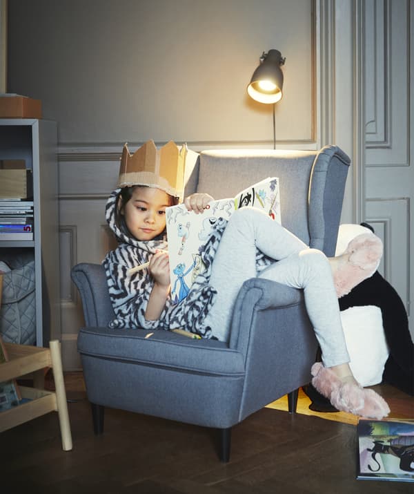 Une petite fille qui porte une couronne en carton et colorie installée dans un fauteuil gris.