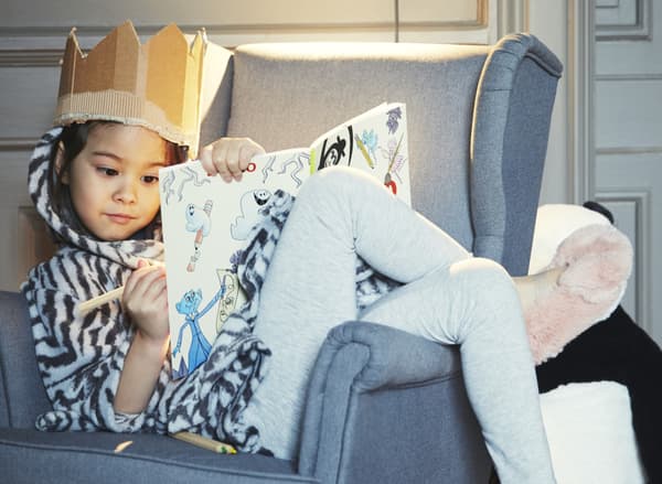 Une petite fille se détend chez elle avec une couronne en papier sur la tête et se prélasse dans un fauteuil tout en dessinant.