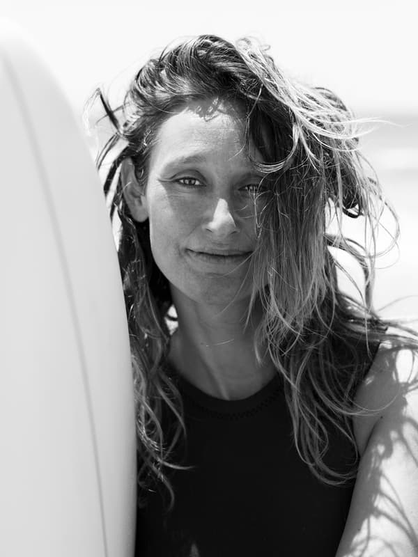 Une photo en noir et blanc de la surfeuse professionnelle retraitée Kassia Meador debout près d’une planche de surf blanche au bord de l’océan.