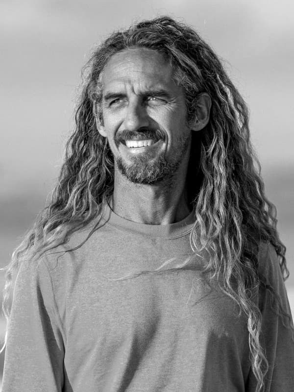Une photo en noir et blanc du surfeur professionnel retraité Rob Machado qui porte un t-shirt et sourit au bord de l’océan.