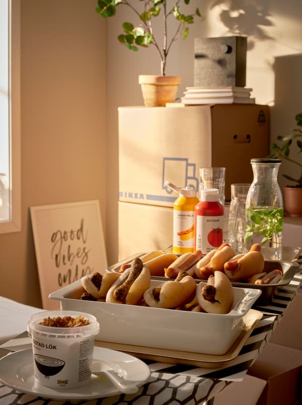 Une pièce baignée de soleil dans laquelle sont empilés des cartons d’emballage JÄTTENE. Un buffet de hot-dogs est servi dans des plats installés sur une table à repasser DÄNKA.