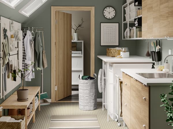 Une pièce spacieuse avec des murs gris-verts et un rangement pour salle de lavage ENHET avec étagères blanches ouvertes et portes et tiroirs effet chêne.
