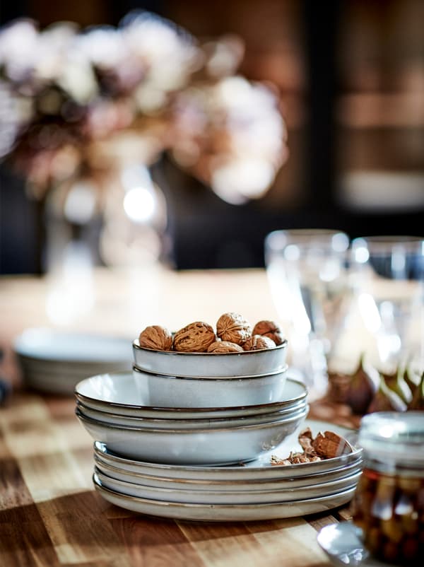 Une pile de bols et assiettes GLADELIG entourée de bocaux, verres et coupe-faim posés sur le plateau en bois d’une table.