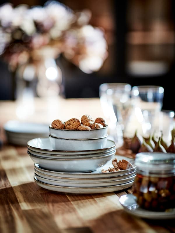 Une pile de bols et assiettes GLADELIG entourée de bocaux, verres et coupe-faim posés sur le plateau en bois d’une table.