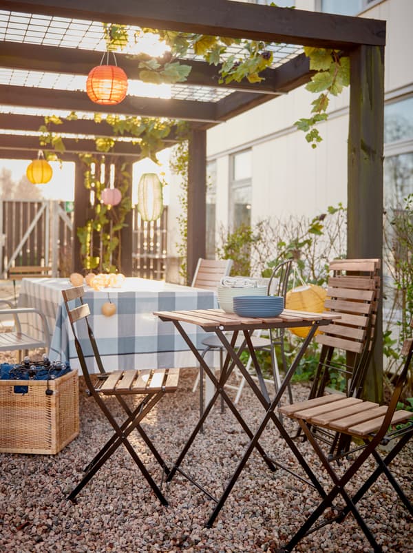Une rangée de chaises et de petites tables en cours d’installation pour une fête sous une pergola dans un jardin.