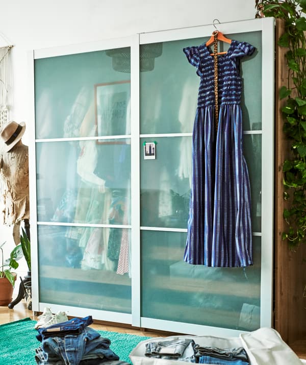 Une robe bleue est suspendue à l’avant d’une garde-robe à portes coulissantes, un mannequin de couture se trouve à côté, et devant, une pile de jeans.