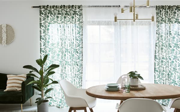 Une salle à manger avec une table en bois et des chaises blanches, devant une porte-fenêtre habillée de rideaux blancs et à motif de feuilles.