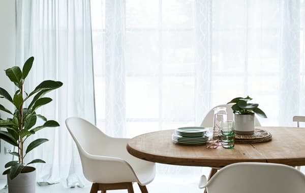 Une salle à manger avec une table en bois ronde et des chaises blanches, devant une porte-fenêtre habillée de rideaux vert et blanc.