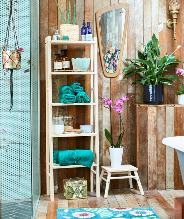 Une salle de bain aux allures de spa, avec une douche à l’italienne carrelée de vert et une étagère en bois garnie de serviettes vertes roulées, de bocaux et de plantes.