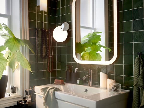 Une salle de bain avec un miroir blanc à éclairage intégré au-dessus d’un meuble lavabo, plus des essuie-mains vert clair et une plante en pot.