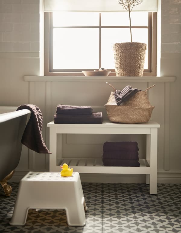 Une salle de bain avec un panier en rotin, un marchepied BOLMEN blanc et un banc HEMNES blanc avec des tablettes pour les serviettes.
