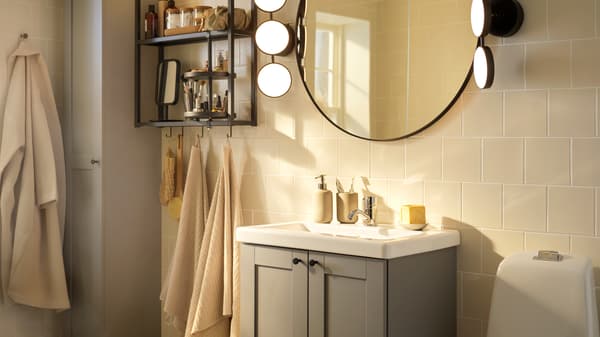 Une salle de bains avec un miroir LINDBYN en noir, des essuie-mains VÅGSJÖN et une robe de chambre BJÄLVEN suspendue près d'un rangement ouvert et fermé.