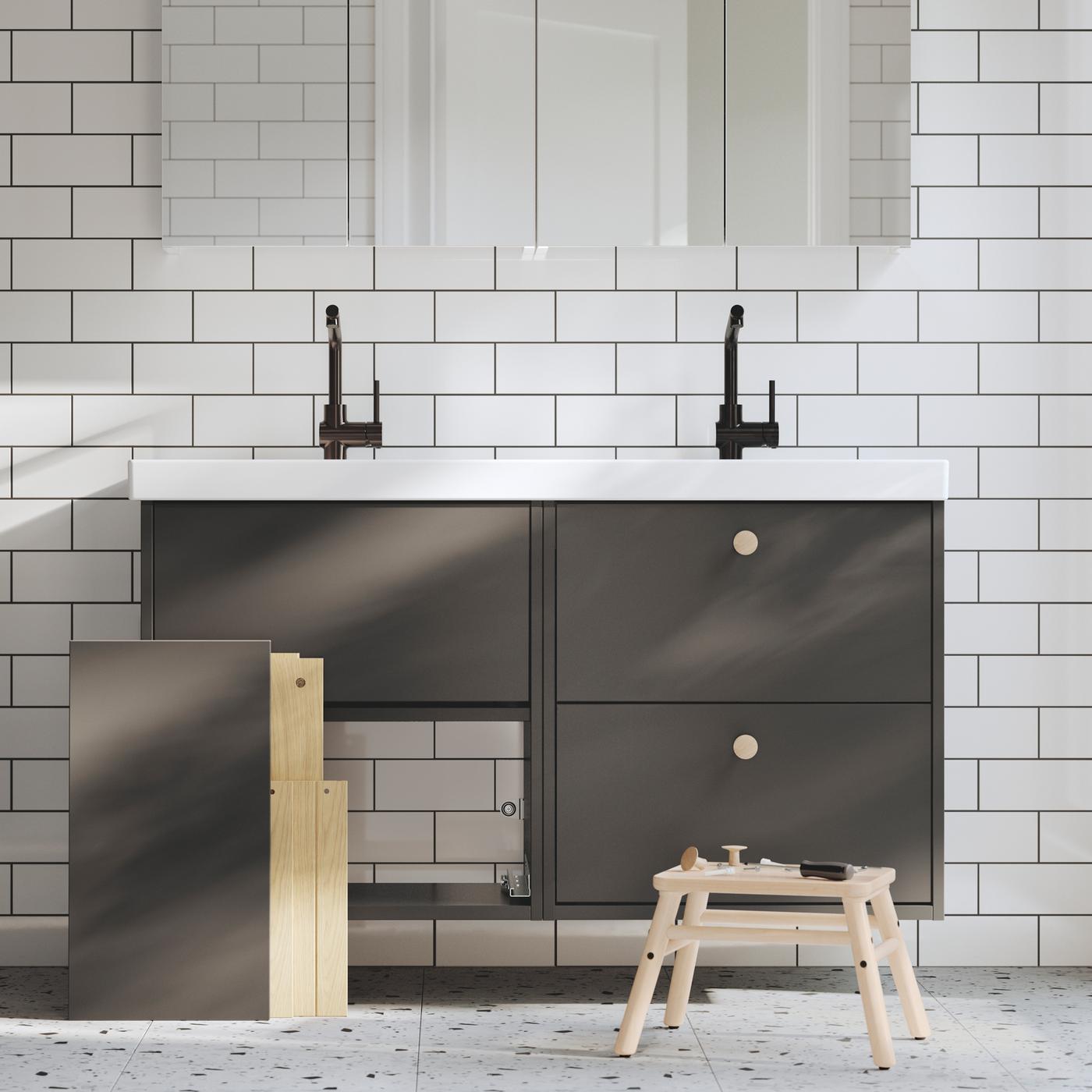 Une salle de bains équipée de deux meubles lavabo HAVBÄCK gris foncé, l’un étant fini et l’autre en cours d’assemblage, avec des outils sur un tabouret.